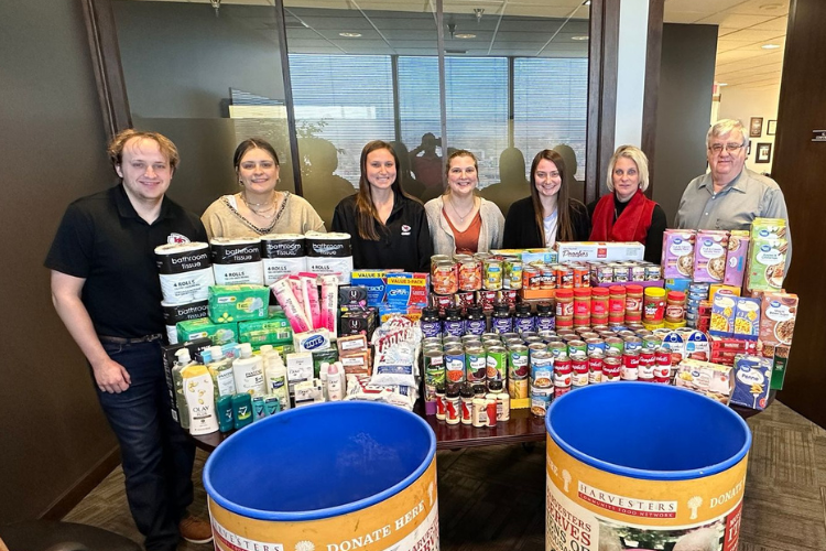 Mize Culture: Team Mize gives back. A group of individuals stand behind cans of food and personal hygiene products collected for Harvesters.